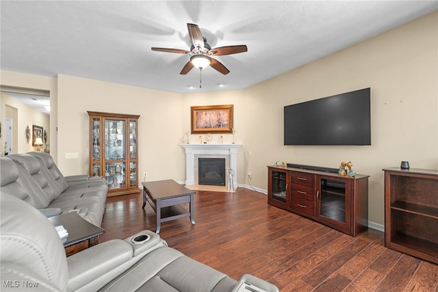 living area featuring baseboards, a fireplace with flush hearth, ceiling fan, and dark wood-style floors