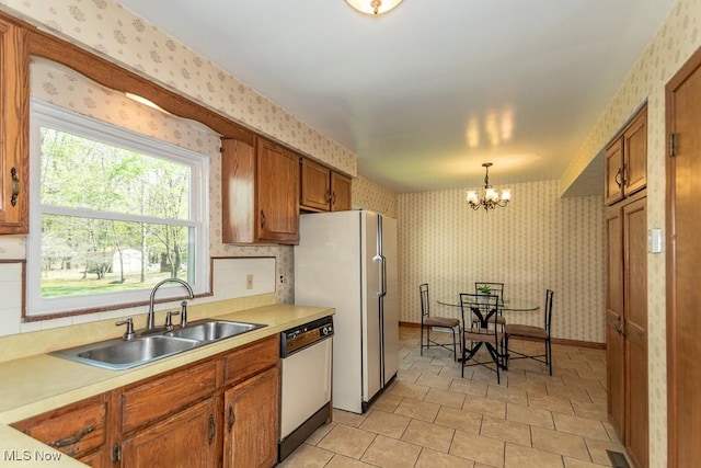 kitchen with white appliances, wallpapered walls, brown cabinets, a sink, and a notable chandelier