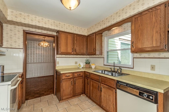 kitchen with brown cabinets, a sink, white appliances, a chandelier, and wallpapered walls