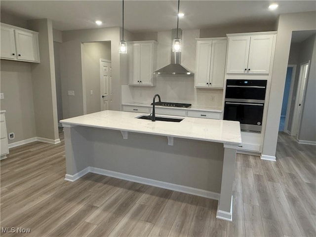 kitchen featuring wall chimney range hood, multiple ovens, a sink, and white cabinetry