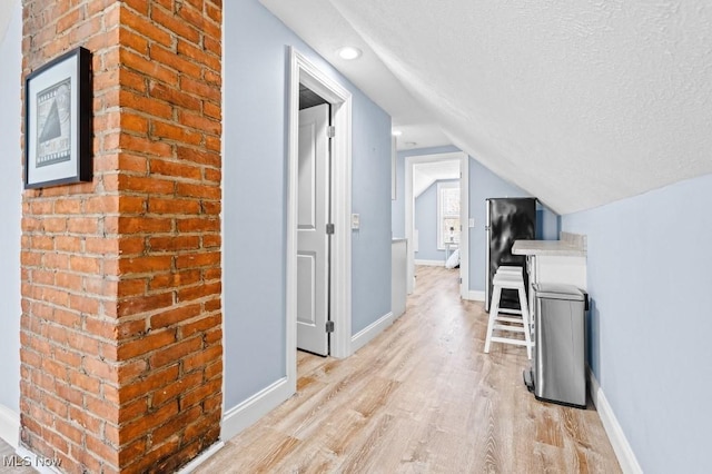 hallway featuring baseboards, light wood-style flooring, vaulted ceiling, and a textured ceiling