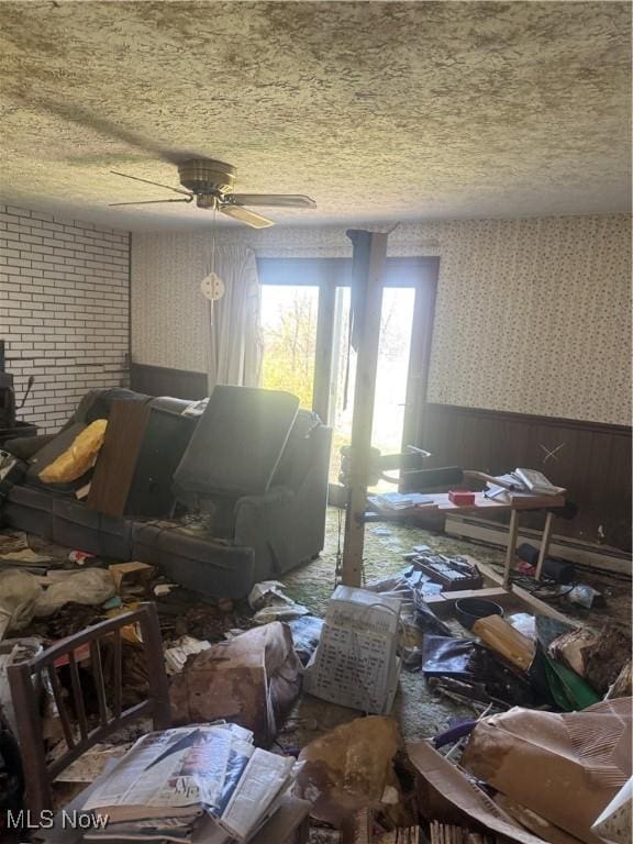 miscellaneous room with a wood stove, wallpapered walls, a textured ceiling, a wainscoted wall, and a ceiling fan