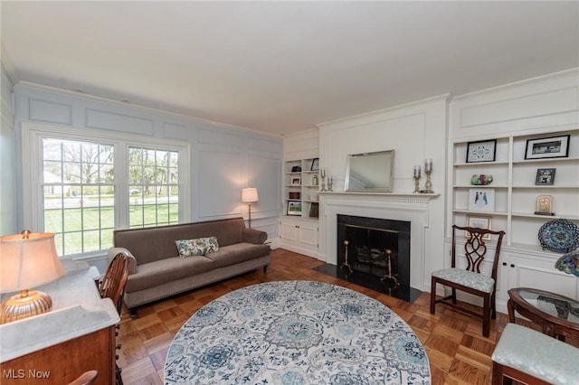 living area with crown molding, a fireplace with flush hearth, a decorative wall, and built in shelves