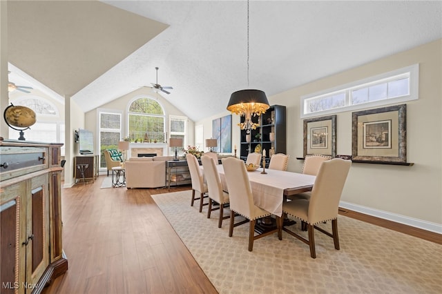 dining room featuring a fireplace, wood finished floors, baseboards, ceiling fan with notable chandelier, and lofted ceiling