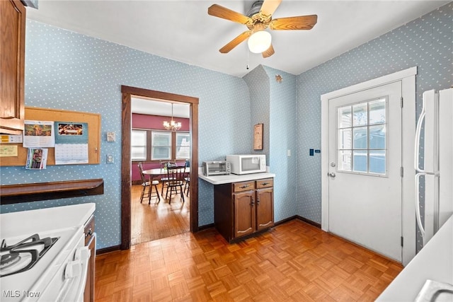 kitchen featuring brown cabinetry, light countertops, ceiling fan with notable chandelier, wallpapered walls, and white appliances