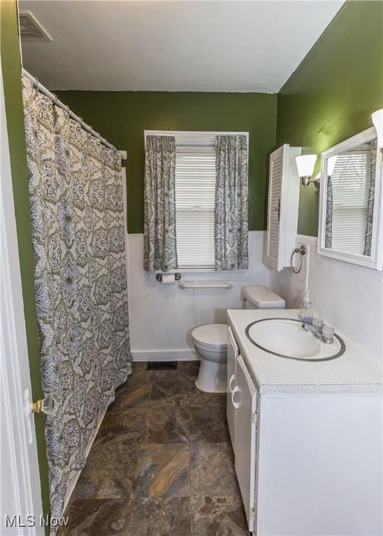bathroom featuring stone finish flooring, visible vents, a wainscoted wall, vanity, and toilet