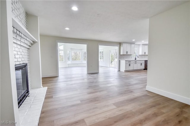 unfurnished living room featuring a brick fireplace, a sink, baseboards, light wood finished floors, and recessed lighting
