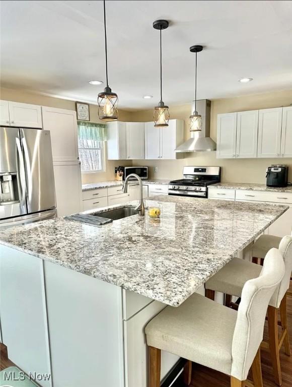 kitchen with a kitchen island with sink, appliances with stainless steel finishes, white cabinetry, a sink, and wall chimney exhaust hood