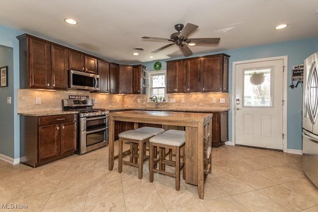 kitchen with decorative backsplash, dark brown cabinetry, ceiling fan, appliances with stainless steel finishes, and a breakfast bar