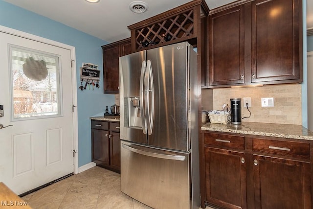 kitchen with dark brown cabinetry, light stone counters, stainless steel fridge, decorative backsplash, and visible vents