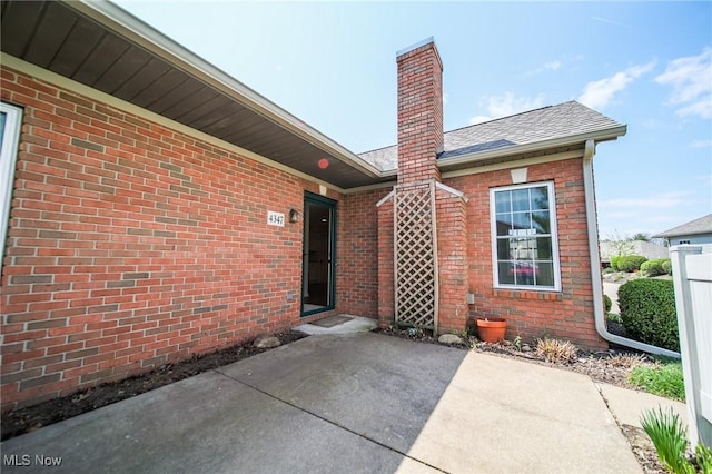 view of exterior entry featuring brick siding, a chimney, a shingled roof, and a patio area