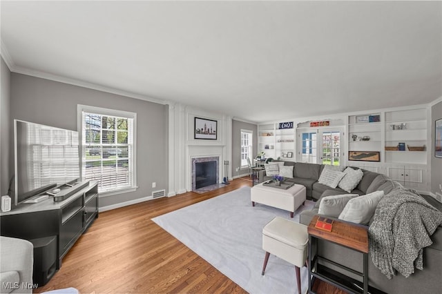 living room featuring light wood-style floors, built in shelves, baseboards, a fireplace with flush hearth, and ornamental molding