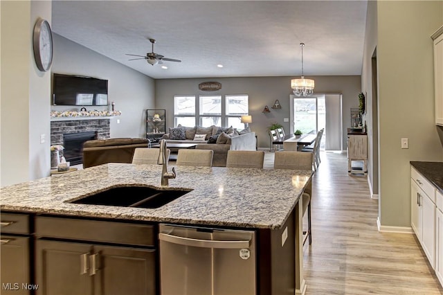 kitchen with stainless steel dishwasher, a fireplace, a sink, light wood finished floors, and ceiling fan with notable chandelier
