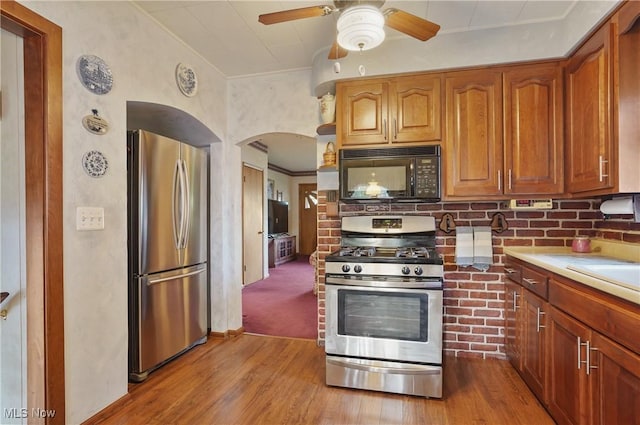 kitchen featuring ceiling fan, brown cabinetry, arched walkways, wood finished floors, and stainless steel appliances