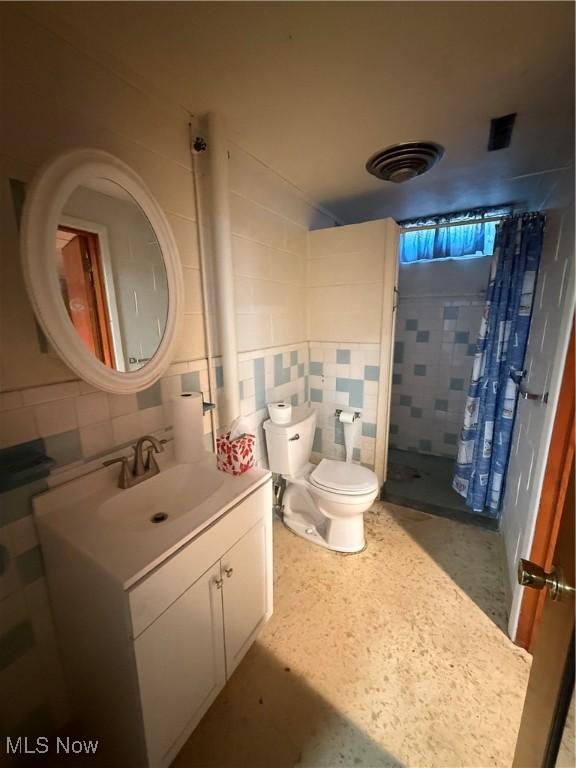 bathroom featuring vanity, a tile shower, toilet, and tile walls
