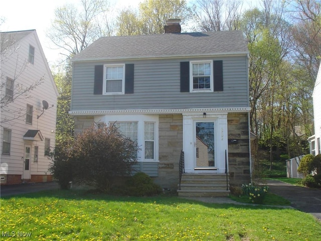 colonial-style house with roof with shingles, a chimney, a front yard, and stone siding