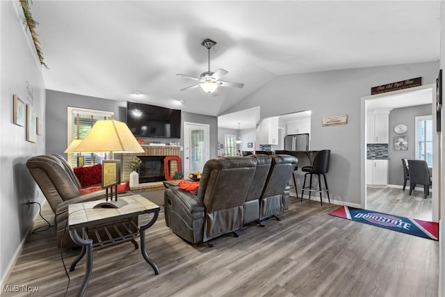 living room with a brick fireplace, a ceiling fan, baseboards, wood finished floors, and vaulted ceiling