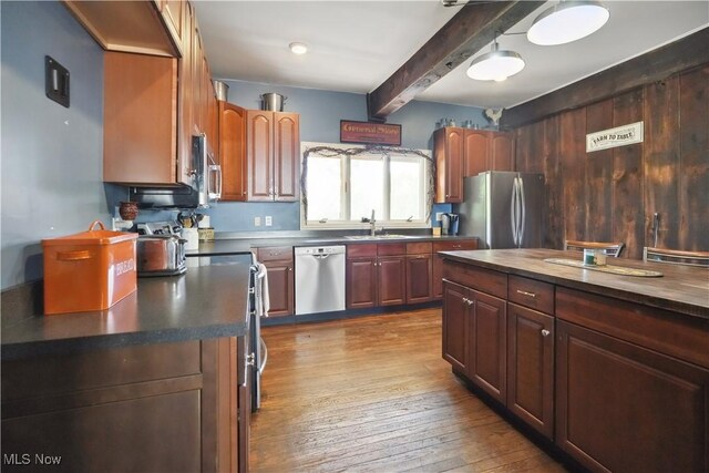 kitchen with appliances with stainless steel finishes, dark countertops, beam ceiling, wood finished floors, and a sink