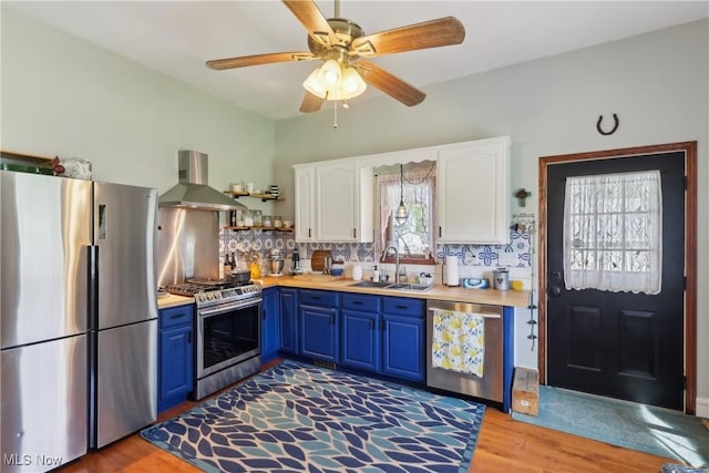 kitchen with wall chimney range hood, stainless steel appliances, a sink, light wood-style flooring, and blue cabinetry