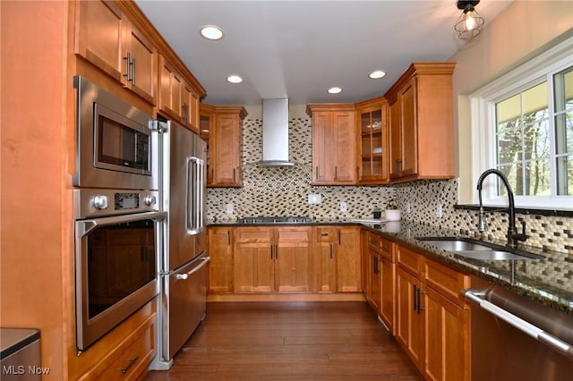 kitchen featuring brown cabinetry, dark stone counters, wall chimney exhaust hood, a sink, and stainless steel appliances
