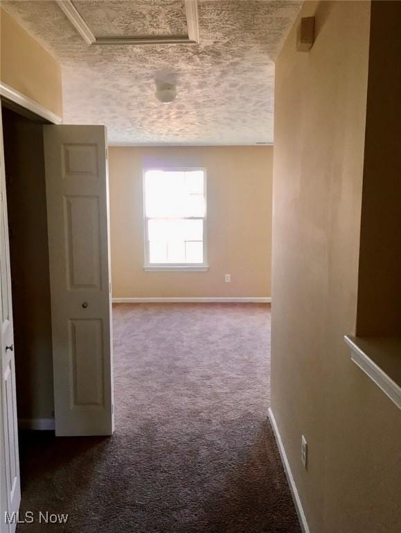 hall with dark carpet, a textured ceiling, baseboards, and attic access