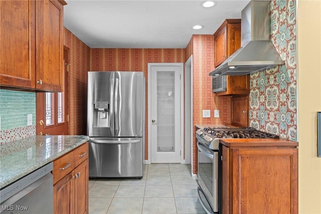 kitchen with light tile patterned flooring, brown cabinetry, wall chimney exhaust hood, stainless steel appliances, and tasteful backsplash