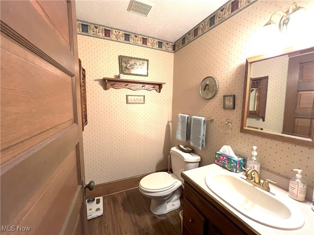 half bath with vanity, toilet, a textured ceiling, wood finished floors, and visible vents
