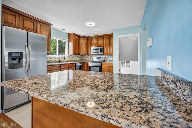 kitchen with decorative backsplash, appliances with stainless steel finishes, hanging light fixtures, brown cabinets, and light stone counters