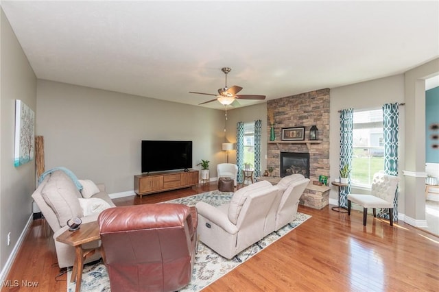 living room featuring a stone fireplace, a ceiling fan, baseboards, and wood finished floors