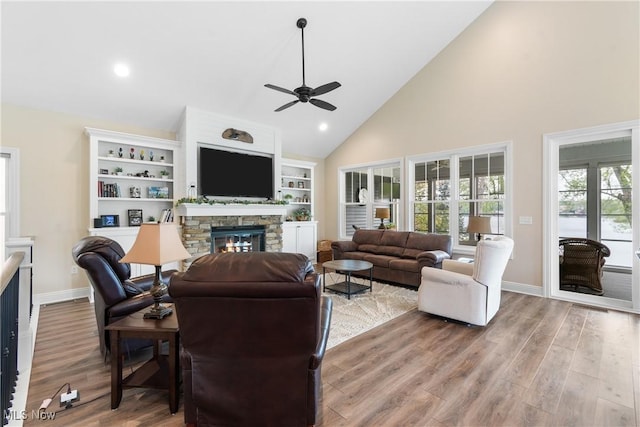 living room with ceiling fan, a stone fireplace, baseboards, high vaulted ceiling, and light wood-style floors
