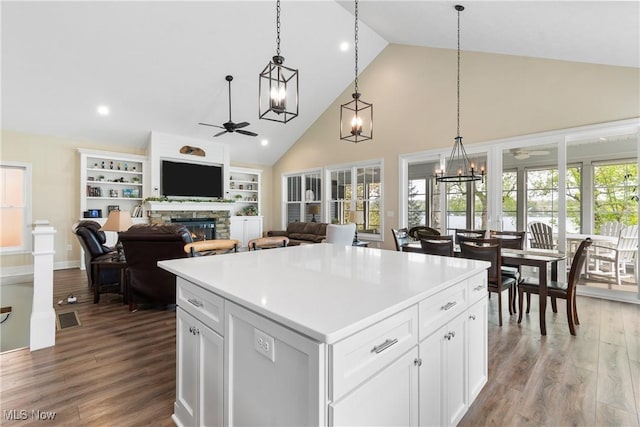 kitchen featuring a stone fireplace, a center island, wood finished floors, high vaulted ceiling, and ceiling fan with notable chandelier