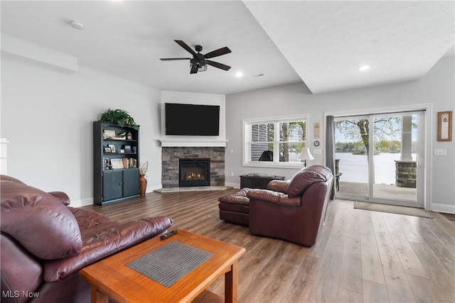 living area featuring baseboards, ceiling fan, a stone fireplace, and wood finished floors