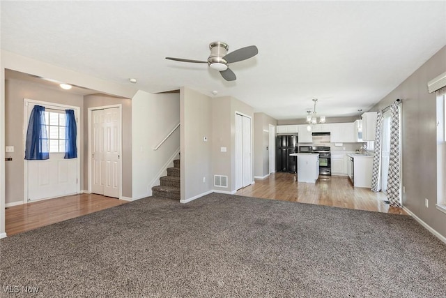 unfurnished living room featuring stairway, baseboards, ceiling fan with notable chandelier, and light colored carpet