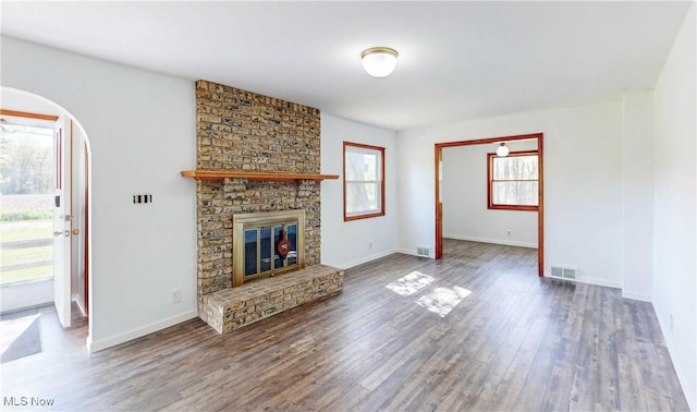 unfurnished living room with a brick fireplace, arched walkways, baseboards, wood finished floors, and visible vents