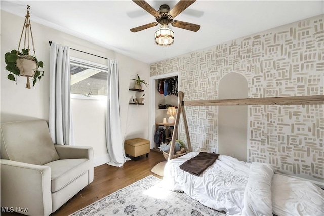 bedroom with ceiling fan, an accent wall, baseboards, wallpapered walls, and wood finished floors