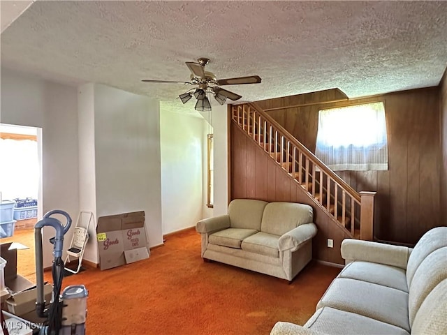 carpeted living room featuring a ceiling fan, a textured ceiling, wooden walls, and stairway