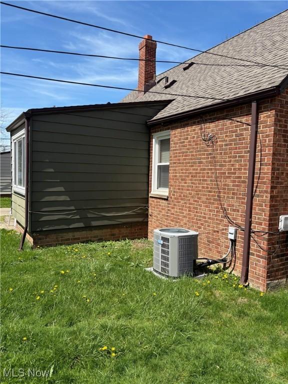 view of home's exterior featuring a chimney, roof with shingles, central AC unit, brick siding, and a yard