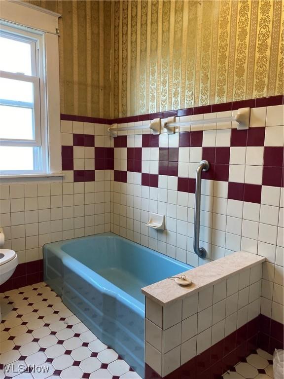 full bath featuring a tub to relax in, wallpapered walls, a wainscoted wall, and tile walls