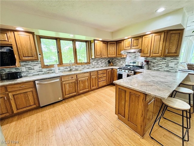 kitchen featuring a breakfast bar area, brown cabinets, a sink, under cabinet range hood, and stainless steel appliances