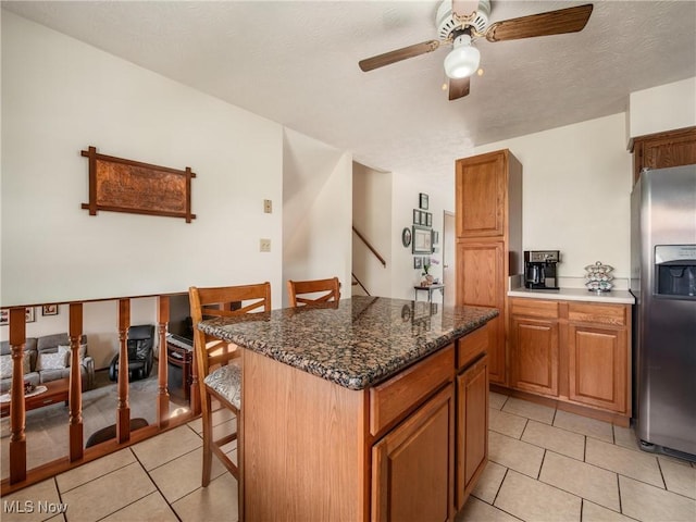 kitchen with brown cabinetry, light tile patterned flooring, ceiling fan, a kitchen breakfast bar, and stainless steel refrigerator with ice dispenser