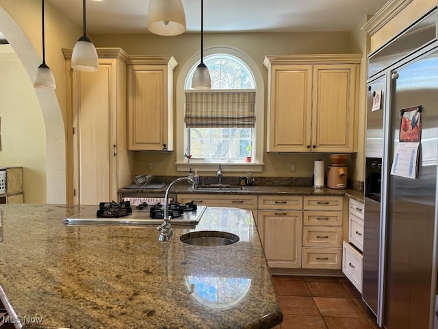 kitchen with a sink, arched walkways, built in fridge, dark stone counters, and decorative light fixtures