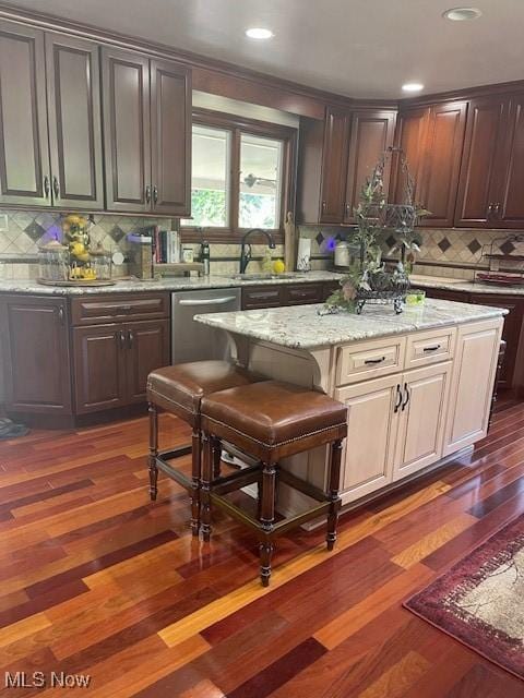 kitchen featuring dishwasher, dark wood-type flooring, a sink, a kitchen island, and backsplash