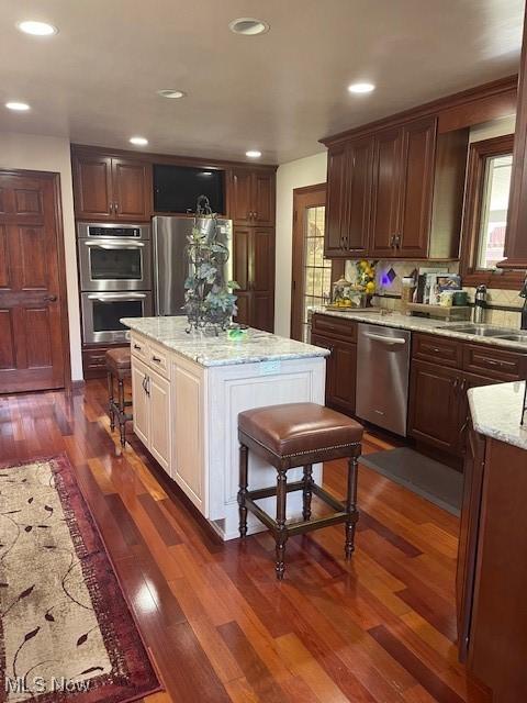 kitchen featuring a sink, appliances with stainless steel finishes, a kitchen island, light stone counters, and dark wood-style floors