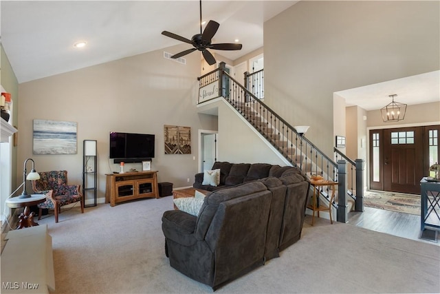 carpeted living area featuring baseboards, visible vents, ceiling fan with notable chandelier, stairs, and high vaulted ceiling