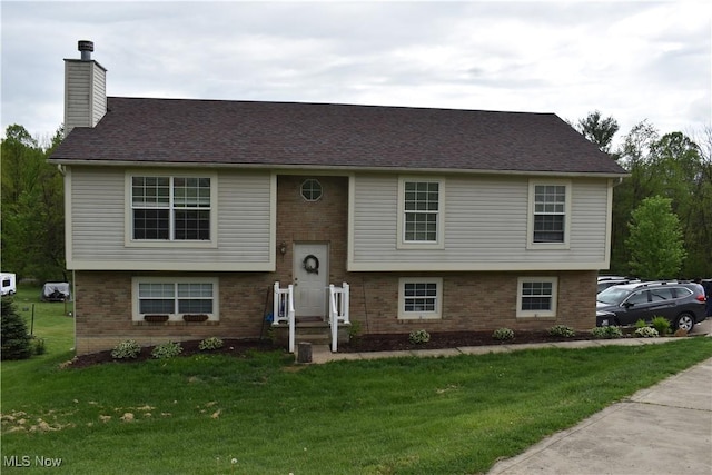 raised ranch with a chimney, a shingled roof, brick siding, and a front lawn