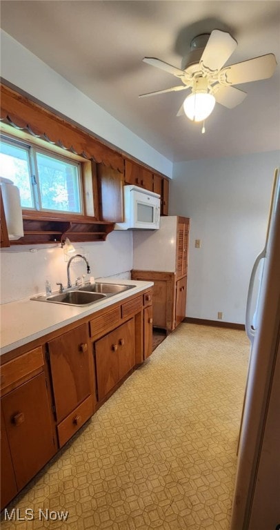 kitchen with a ceiling fan, a sink, brown cabinetry, white appliances, and light countertops