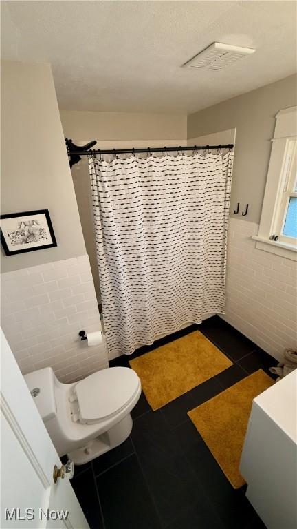 full bath featuring toilet, a shower with curtain, tile patterned floors, tile walls, and wainscoting