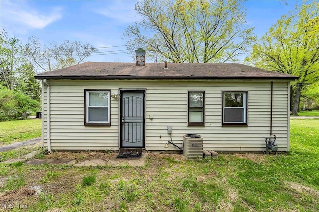 back of property featuring a chimney, central air condition unit, a shingled roof, and a lawn