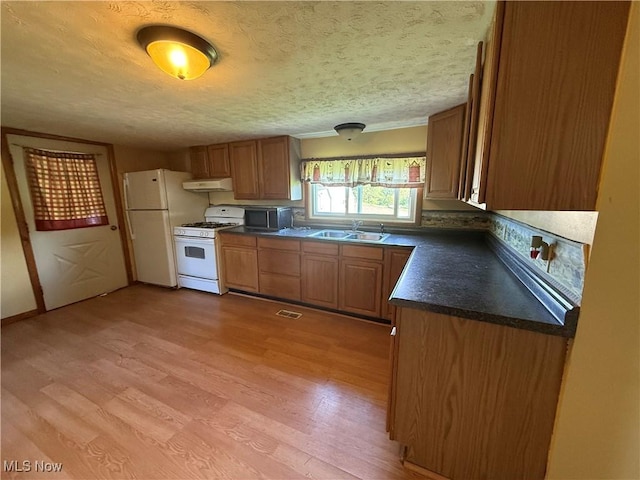 kitchen with a sink, white appliances, under cabinet range hood, light wood finished floors, and dark countertops