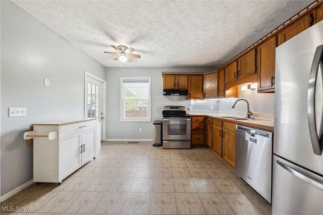 kitchen featuring stainless steel appliances, under cabinet range hood, a ceiling fan, baseboards, and brown cabinets